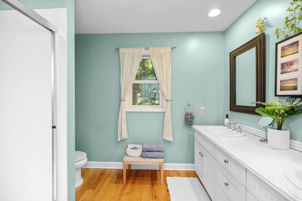 Light and airy bathroom with light blue-green walls and hardwood floors, featuring a toilet and a window with sheer white curtains. A small wooden bench with folded towels is placed beneath the window.