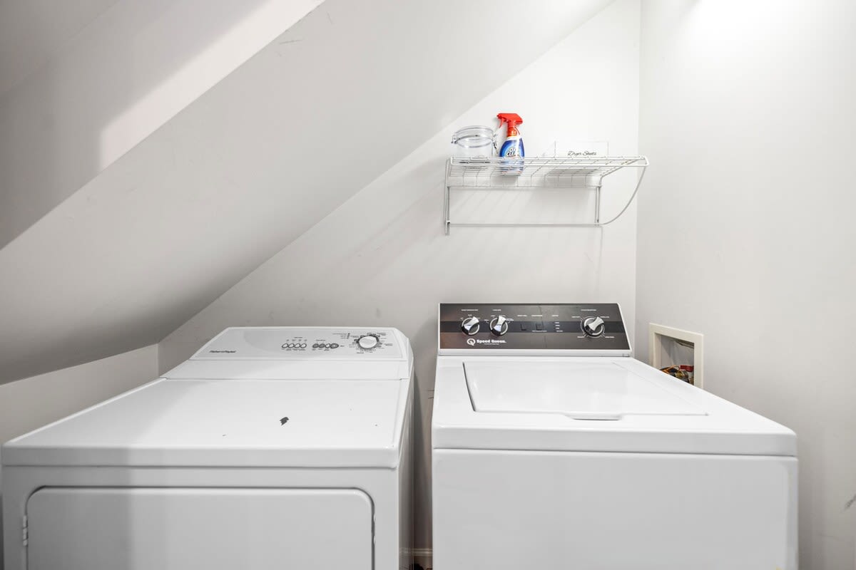 Compact laundry area with white walls and a sloped ceiling, housing a white top-loading washing machine and a matching dryer. A wire shelf above provides storage for laundry essentials.
