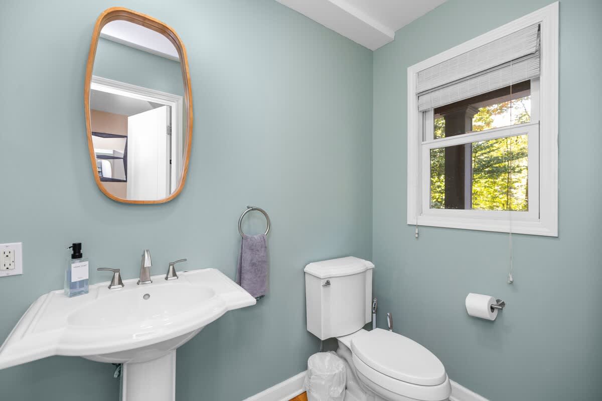 Powder room with light blue-green walls, a white pedestal sink, and a toilet. A wooden mirror hangs above the sink, and a window with blinds offers natural light.