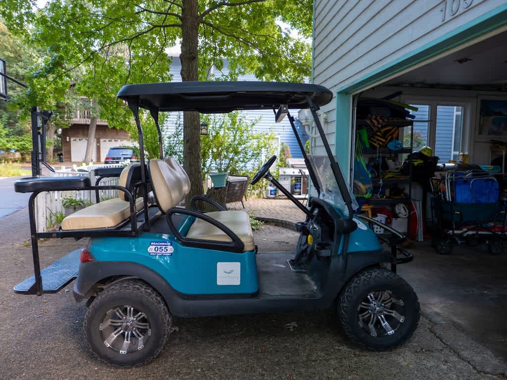 A teal golf cart with tan seats is parked inside a garage or carport area, with various outdoor equipment visible in the background and a white house exterior.