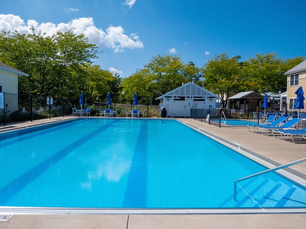 Large outdoor swimming pool with clear blue water, surrounded by blue lounge chairs and umbrellas, with a white pool house and lush green trees under a bright blue sky.
