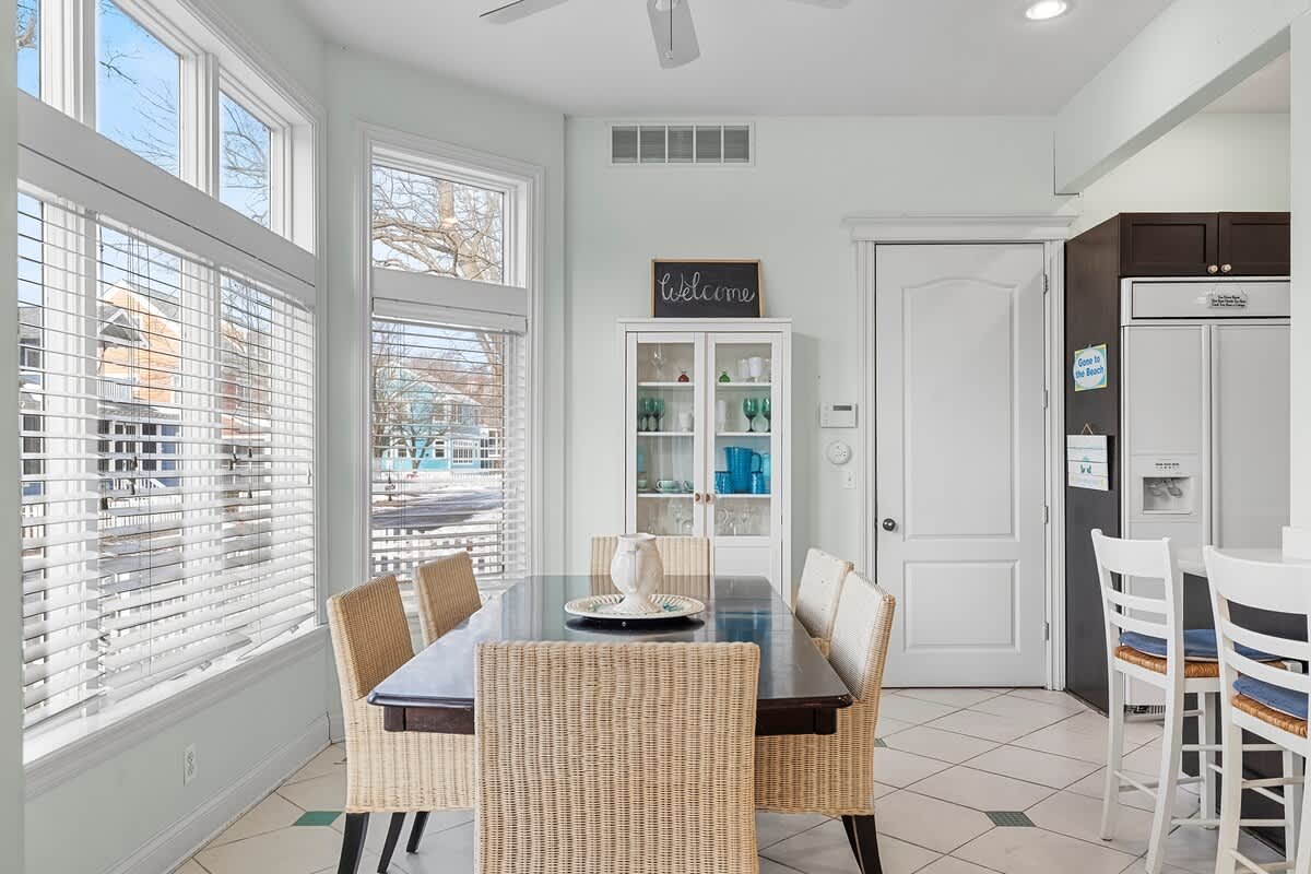 Bright dining area featuring a dark wood table surrounded by wicker chairs, positioned next to large windows with white blinds, and a white display hutch with a "welcome" sign.