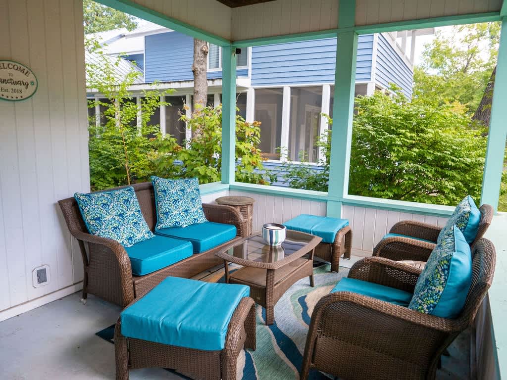 A cozy covered porch featuring wicker outdoor furniture with bright turquoise cushions and patterned pillows, centered around a glass-top coffee table. The space is surrounded by lush greenery, offering a relaxing outdoor retreat.