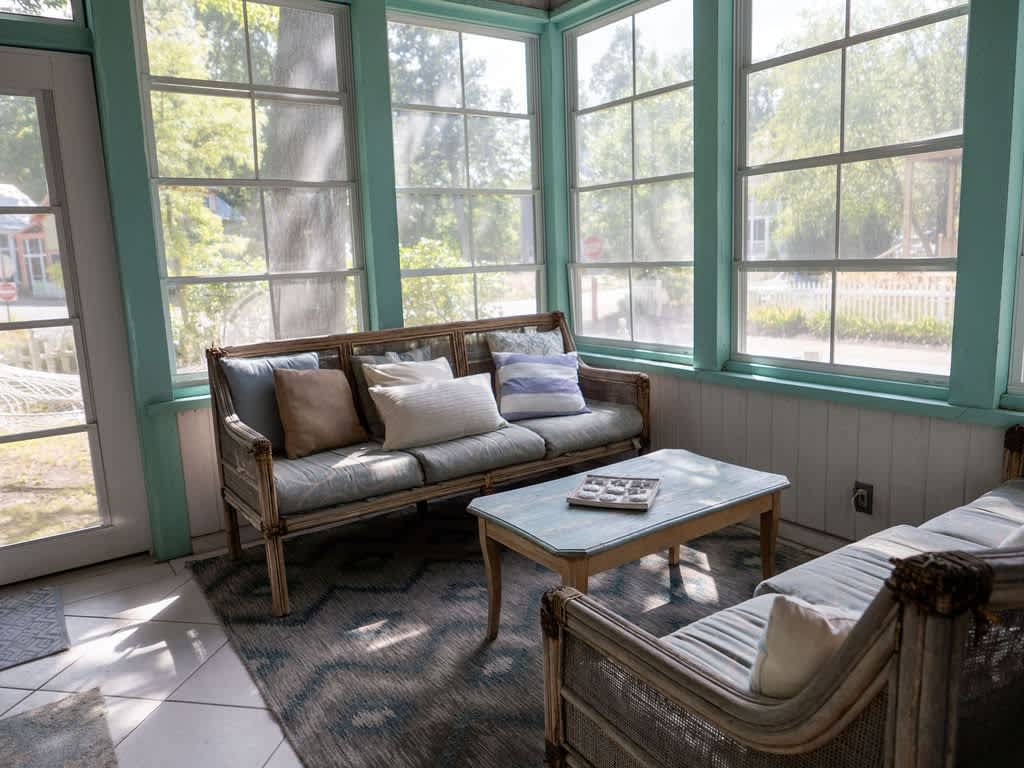 Bright sunroom with large windows and light blue trim, furnished with wicker sofas and a coffee table, featuring light blue cushions and a patterned area rug.