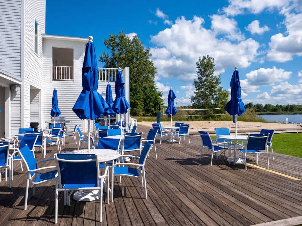 An expansive outdoor deck features numerous white tables and blue chairs, shaded by large blue umbrellas, under a bright blue sky with white clouds. A sandy beach volleyball court and a lake are visible in the background.