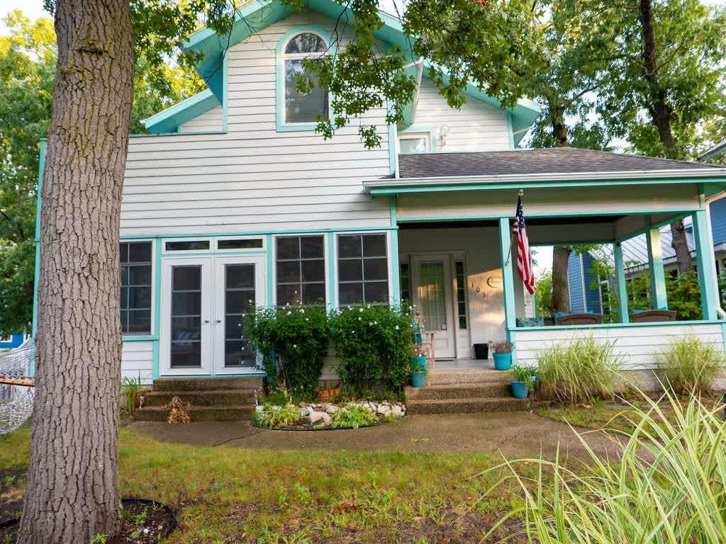 The front exterior of a two-story house with white siding and light teal trim, featuring a covered porch with an American flag, an enclosed sunroom on the left, and mature trees in the foreground and background.