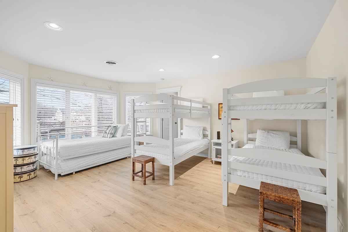 A spacious bedroom with light wood-look flooring and large windows, featuring two white bunk beds, a white daybed with a trundle, and small wooden stools. The room is brightly lit and has a light, airy feel.
