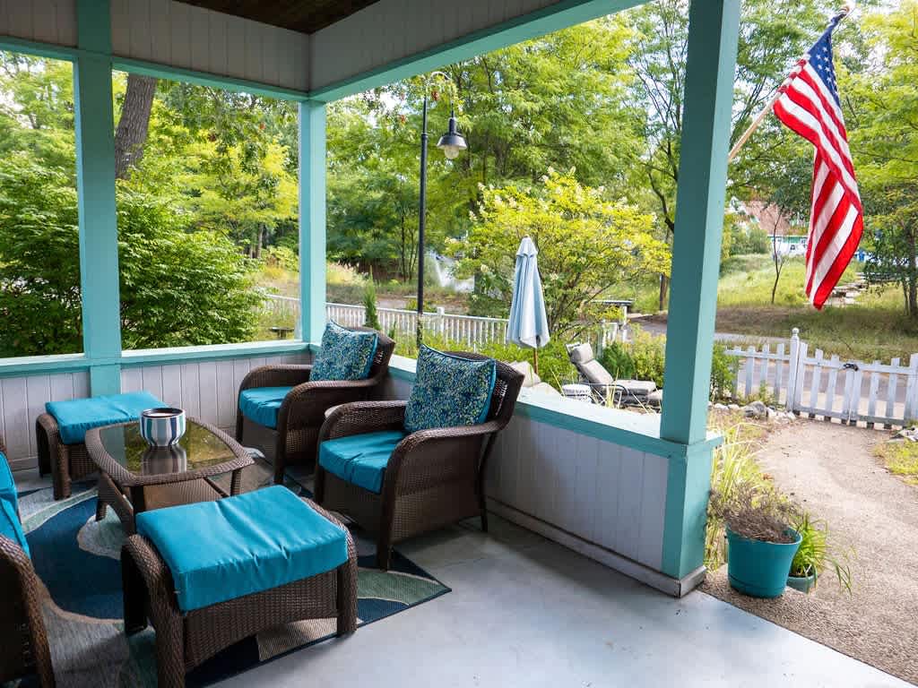 A welcoming covered porch with wicker seating and turquoise cushions, overlooking a green yard with trees and a white picket fence. An American flag hangs from a pole, adding a patriotic touch to the outdoor setting.