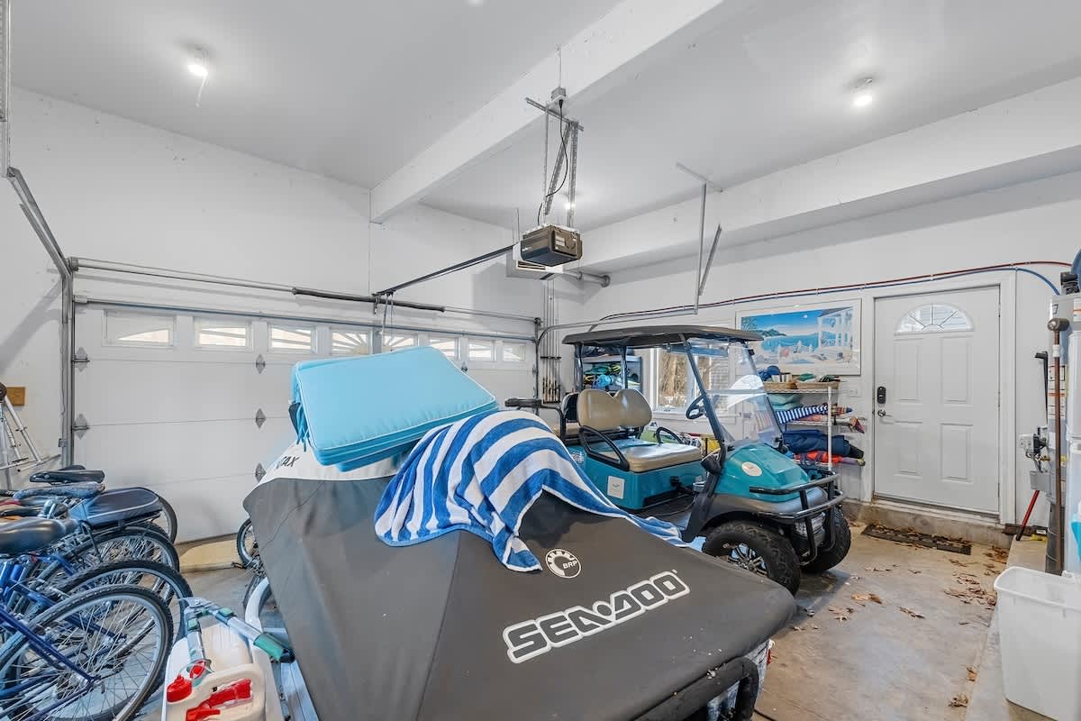 A spacious garage with white walls and exposed ceiling beams, housing a covered jet ski, a golf cart, bicycles, and various storage items. The garage door is visible, along with a side entry door.