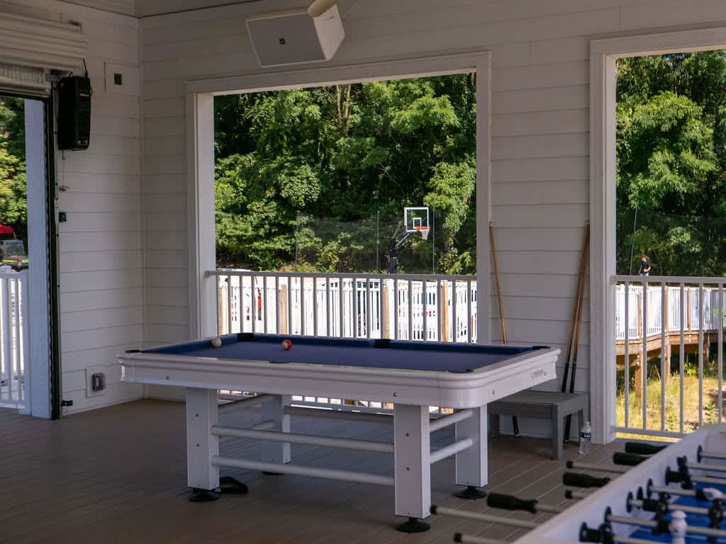 A close-up view of a blue-felt pool table with a white frame, set on light composite decking in an enclosed porch. Beyond the railing, a lush green forest and a basketball hoop are visible through large open windows.