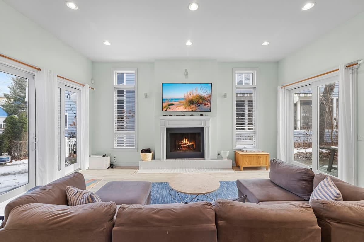 Spacious living room centered around a white fireplace with a wall-mounted TV above it, featuring a large brown sectional sofa, a round coffee table, and multiple French doors leading to the exterior.