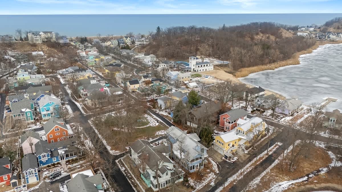 An aerial view of a coastal town with colorful houses nestled among trees, leading to a frozen body of water on the right and a vast blue ocean in the distance. The landscape is covered in a light dusting of snow.