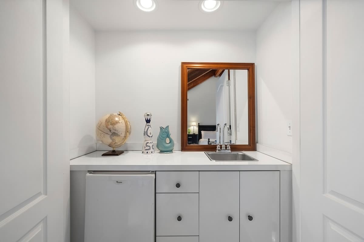 Compact wet bar area with white cabinetry, a mini-fridge, a stainless steel sink, and a wooden-framed mirror above the counter. Decorative items like a globe and vases are displayed.
