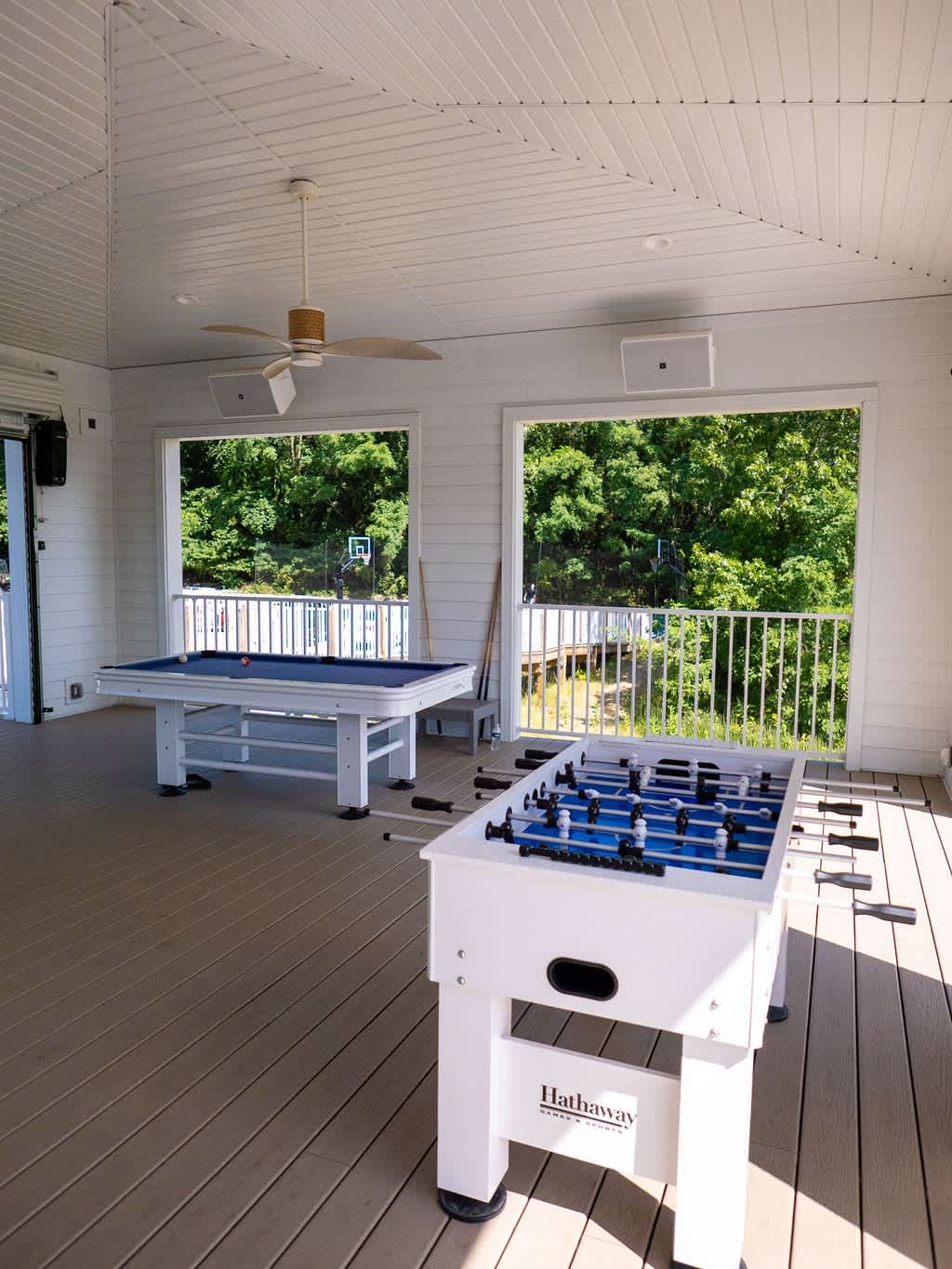 A bright, enclosed game room features a blue-felt pool table and a white foosball table on light wood-look composite decking. Large open windows overlook a lush green forest and a distant basketball hoop.