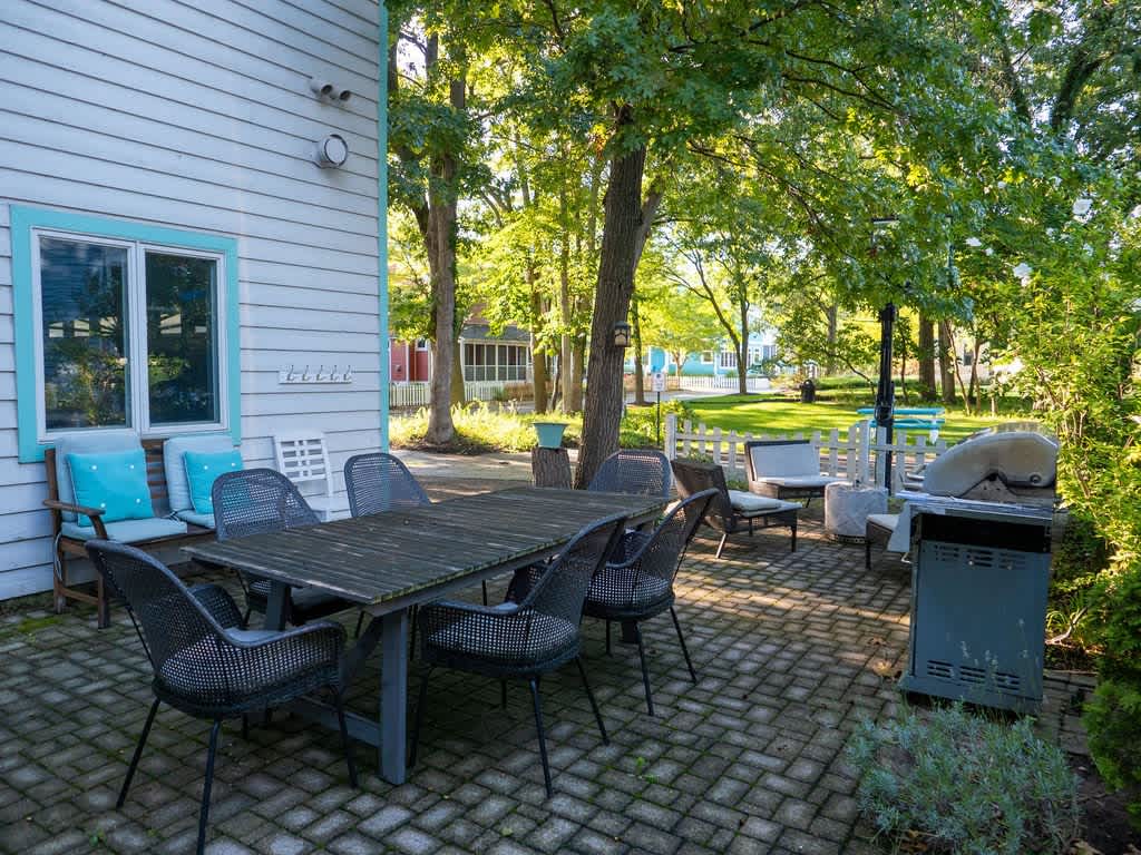 An inviting outdoor patio with a paved surface, featuring a large wooden dining table with several chairs and a BBQ grill. The area is shaded by mature trees and surrounded by lush greenery, perfect for al fresco dining.