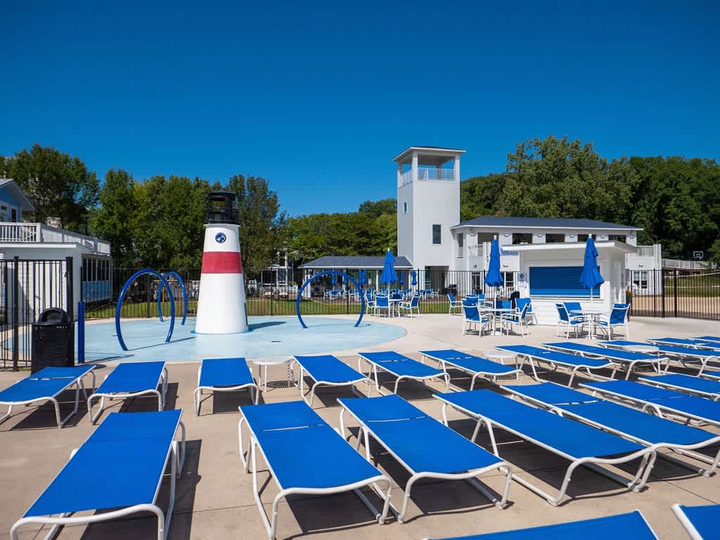 Expansive outdoor pool area featuring numerous blue lounge chairs, a white and red lighthouse-themed splash pad structure, and a modern white building with blue accents in the background.