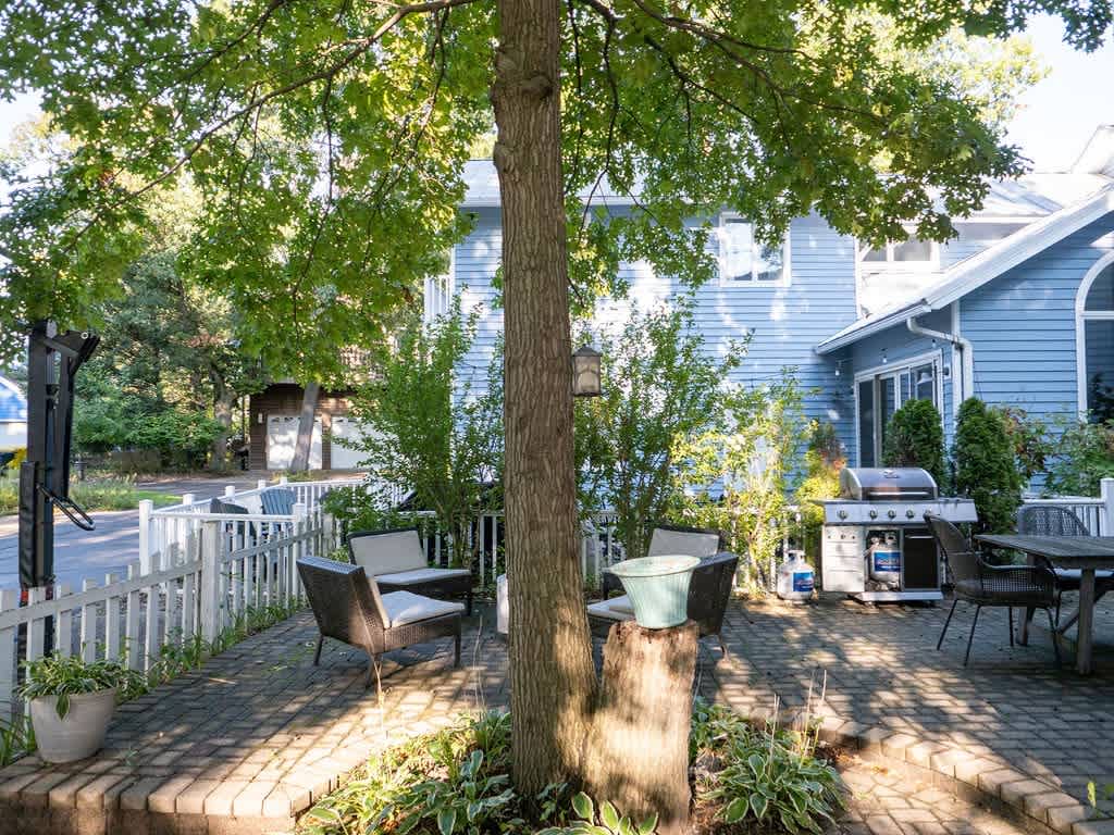 Outdoor brick patio with a large tree providing shade, featuring a seating area with dark chairs and a light blue planter, and a stainless steel BBQ grill next to a light blue house.
