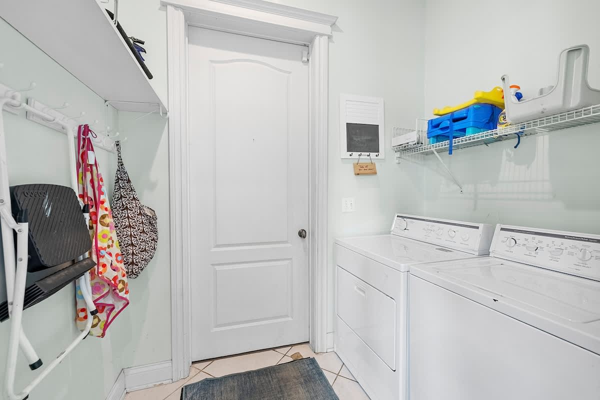 A functional laundry room with light green walls, featuring a white washer and dryer, wire shelving for storage, and a white door. Various laundry supplies and beach towels are visible, indicating practical use.