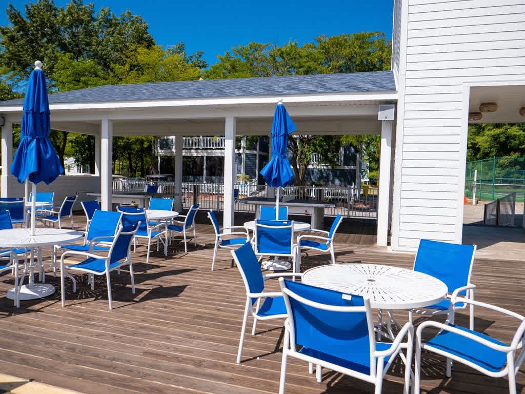 An expansive outdoor deck offers several white round tables with blue chairs and matching umbrellas, providing a comfortable al fresco dining or lounging space adjacent to a white-sided building and surrounded by greenery.