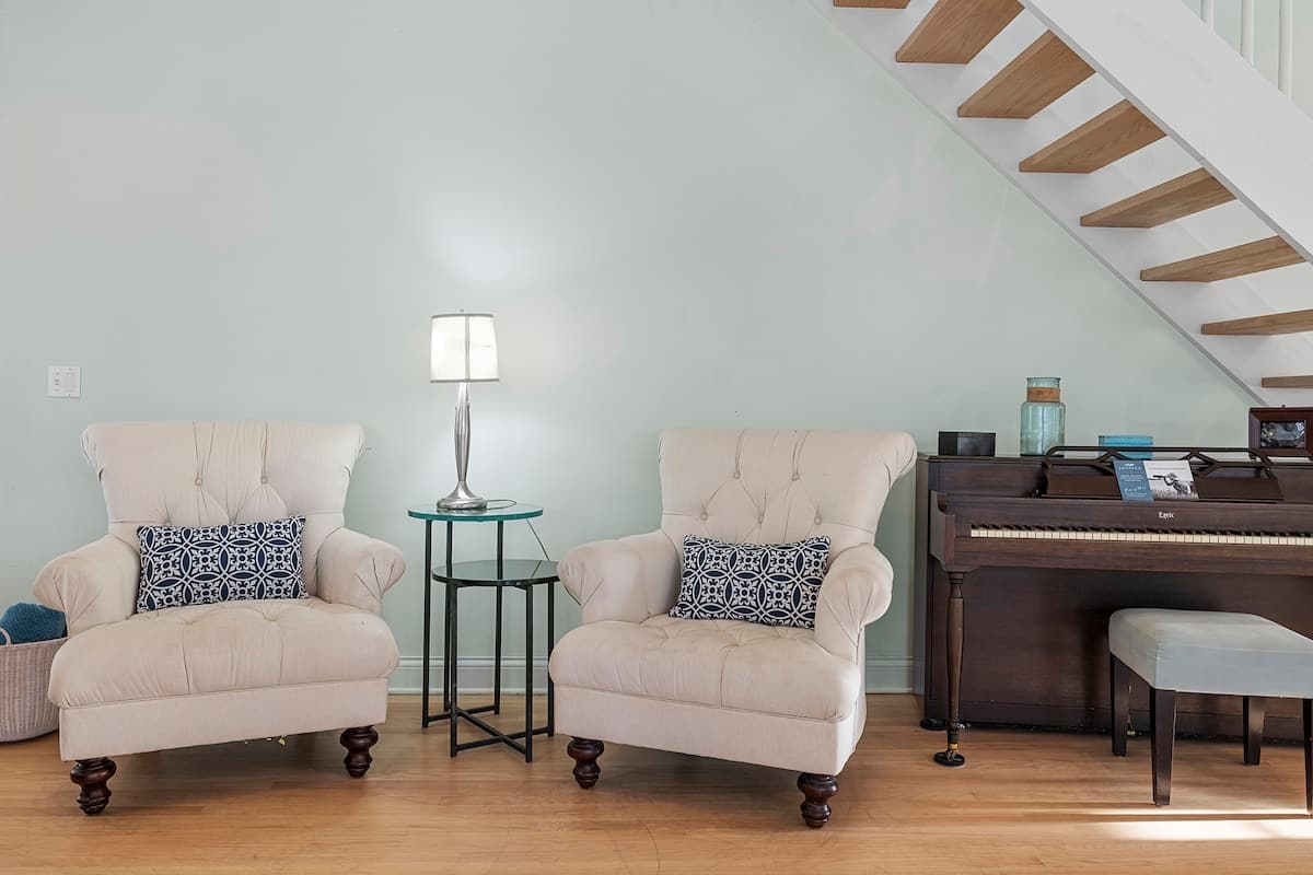 Cozy living space featuring two cream-colored tufted armchairs with patterned throw pillows, a glass-top side table, and a dark wood upright piano, all set against light green walls with a modern staircase in the background.