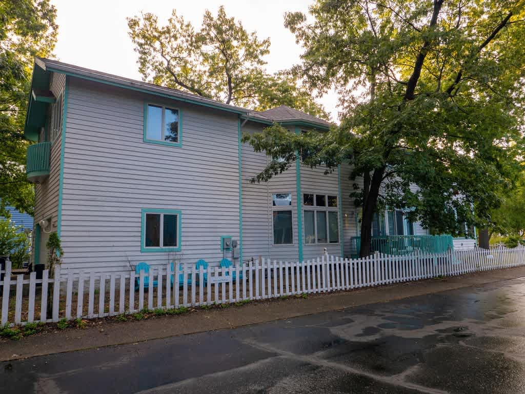 Side exterior view of the white house with light blue trim, featuring multiple windows, a white picket fence along the street, and several light blue Adirondack chairs in the yard.