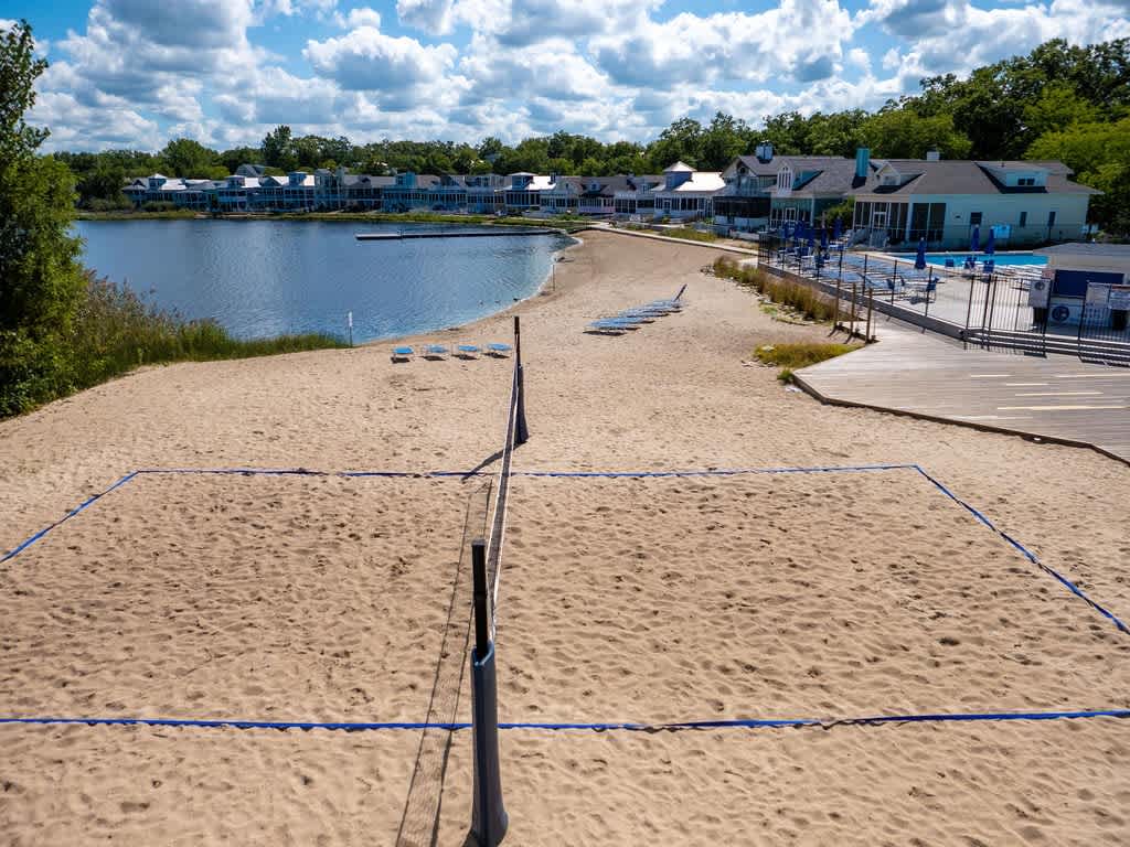 A well-maintained beach volleyball court with a net and boundary lines is set on soft sand, adjacent to a calm lake and a wooden deck with lounge chairs, under a bright sky with scattered clouds.