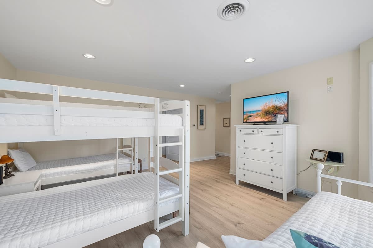 A bright bedroom with light wood-look flooring, featuring white bunk beds with white bedding, a white dresser with a flat-screen TV, and recessed lighting. The room appears spacious and designed for multiple occupants.