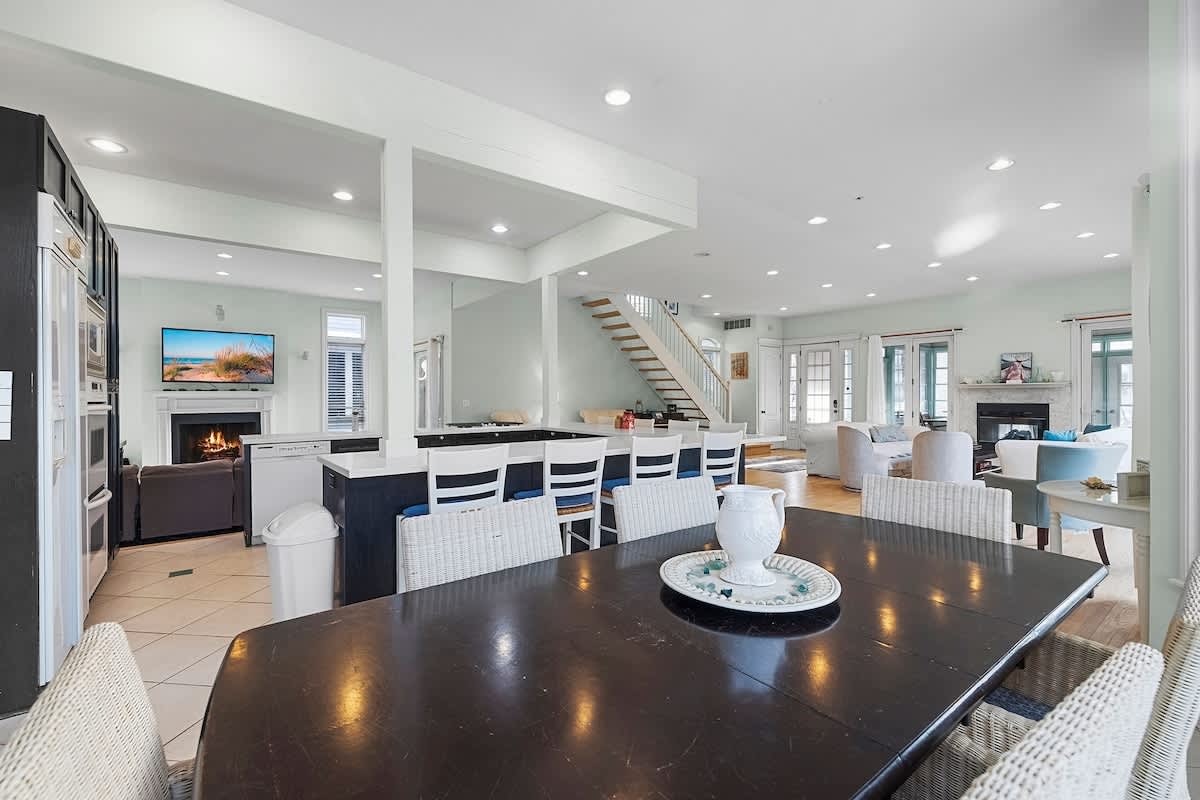 Expansive open-concept space featuring a large kitchen island with bar seating, a dark wood dining table, and a living area with a fireplace and wall-mounted TV, all connected by light green walls and tiled flooring.