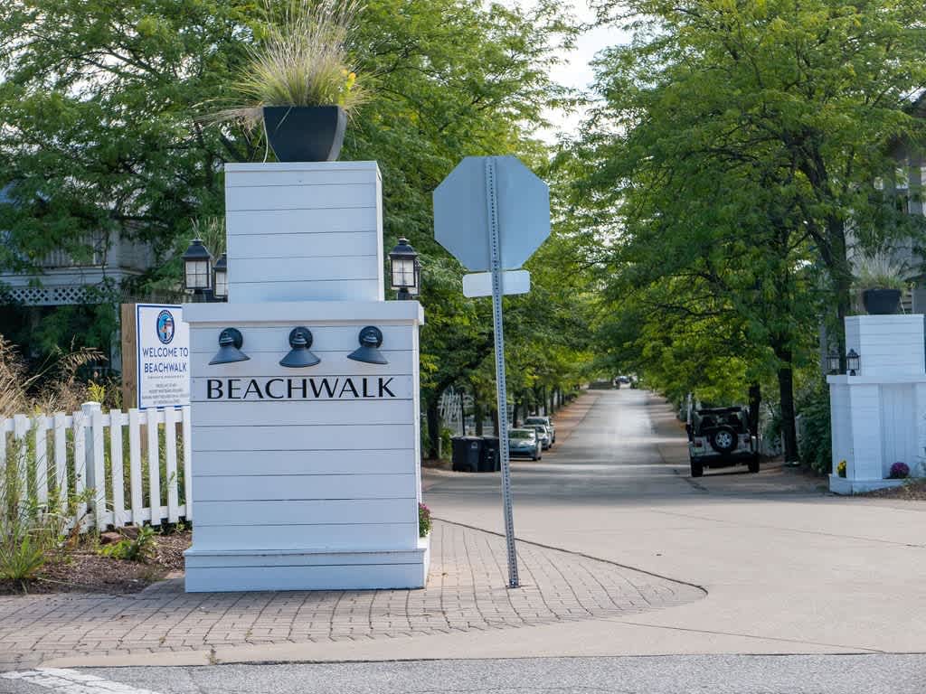 Entrance sign for the 'BEACHWALK' community, a white structure with black lettering and lanterns, marking the paved road leading into the tree-lined neighborhood.
