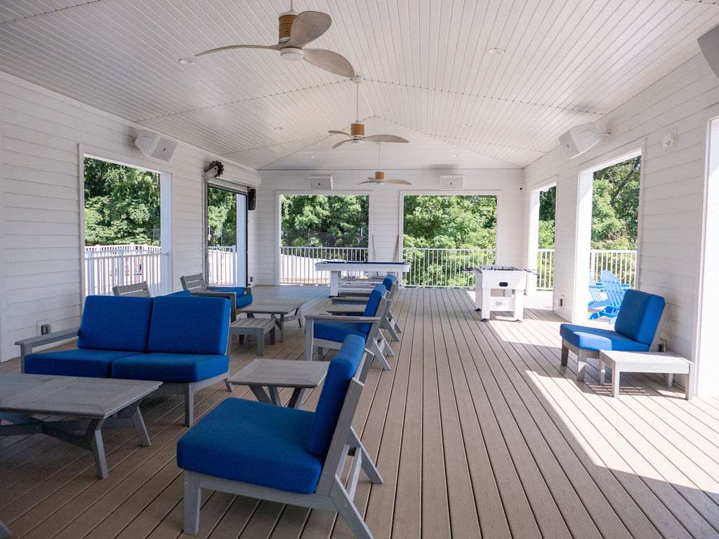 A spacious covered outdoor game room features multiple blue cushioned seating areas, a foosball table, and a pool table, all set on a light wood-toned deck with ceiling fans and views of surrounding greenery.