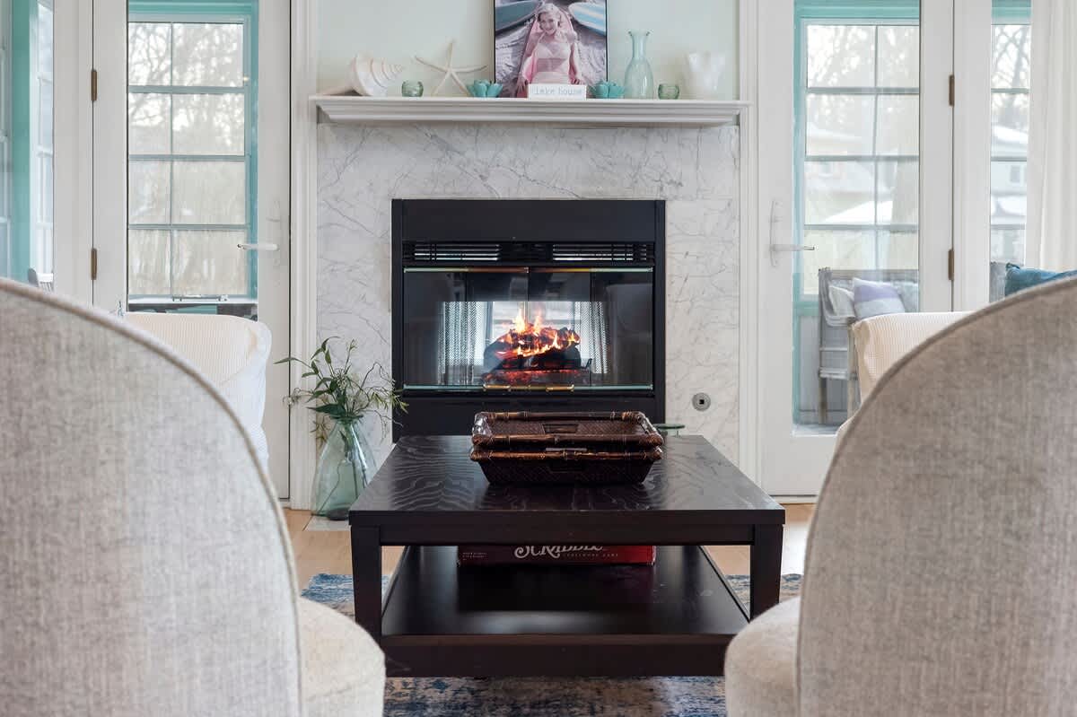 A close-up view of a cozy living room area focusing on a white marble fireplace with a burning fire, flanked by two light gray armchairs and a dark wood coffee table with a decorative box.