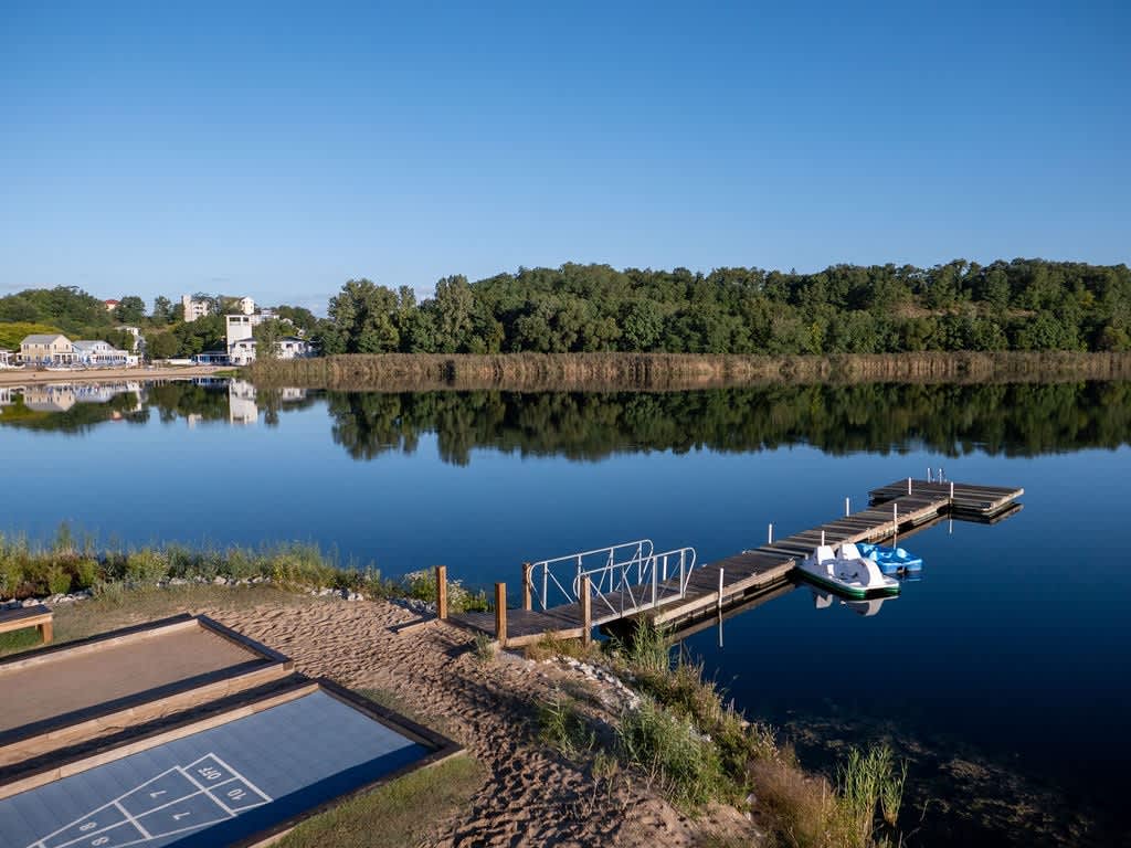 An aerial view captures a serene lake with a wooden dock extending into the water, featuring two paddle boats, alongside a sandy beach area with shuffleboard courts and lush green trees reflecting on the calm surface.