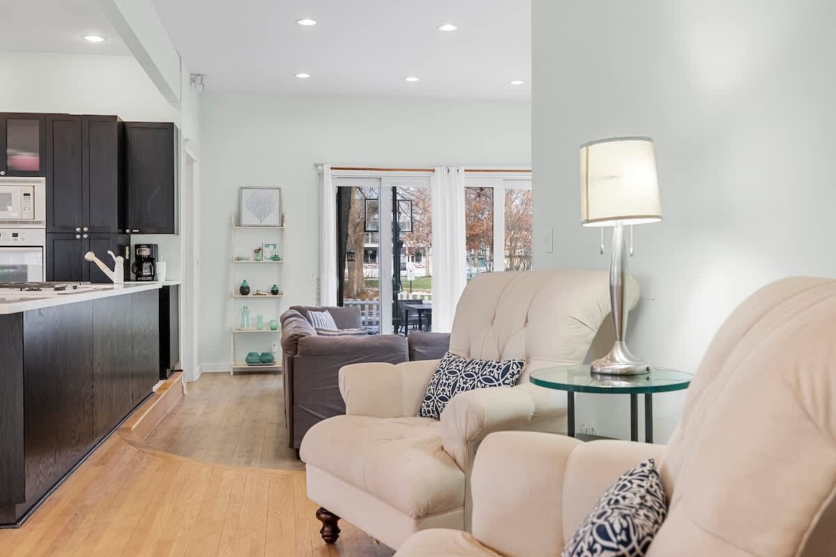 A bright open-concept living space with light green walls and hardwood floors, featuring two light-colored armchairs, a side table with a lamp, and a view towards the kitchen island and dark cabinetry in the background.