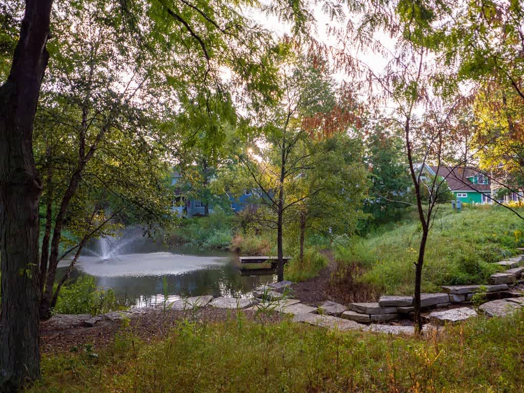 A tranquil pond with a central fountain is nestled within a lush, natural landscape, surrounded by tall trees and green foliage, with a stone path leading down to the water's edge and houses visible in the distance.