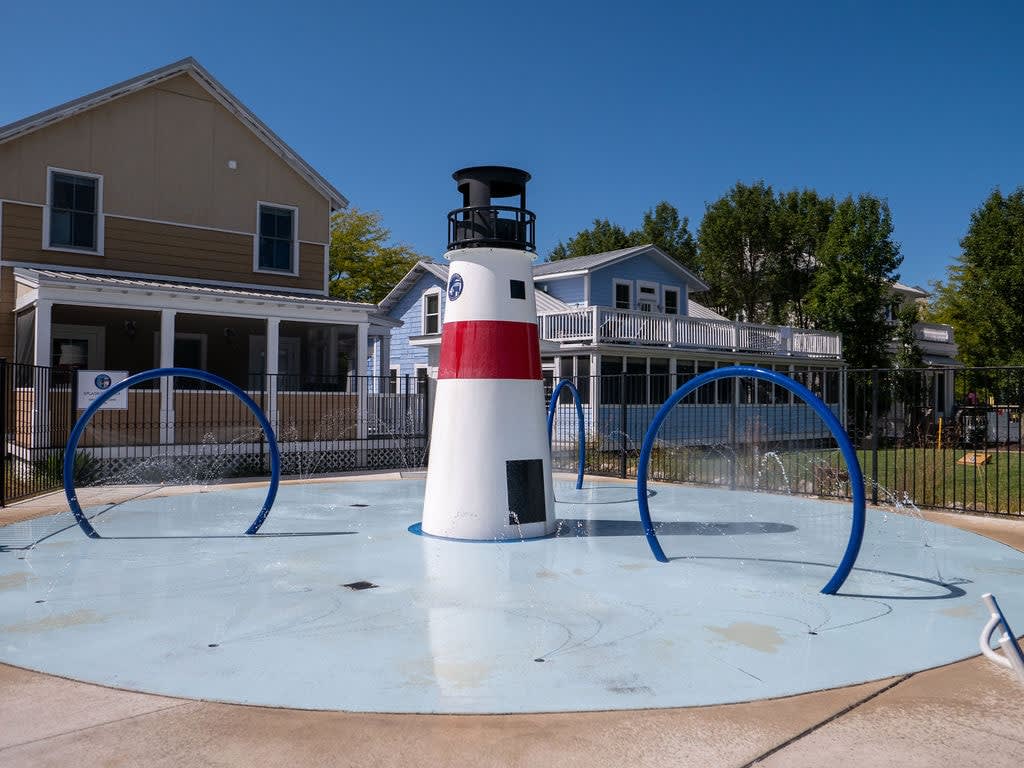 A vibrant splash pad features a white and red striped lighthouse centerpiece, surrounded by blue arched water sprayers on a light blue circular surface, with houses and trees in the background under a clear sky.