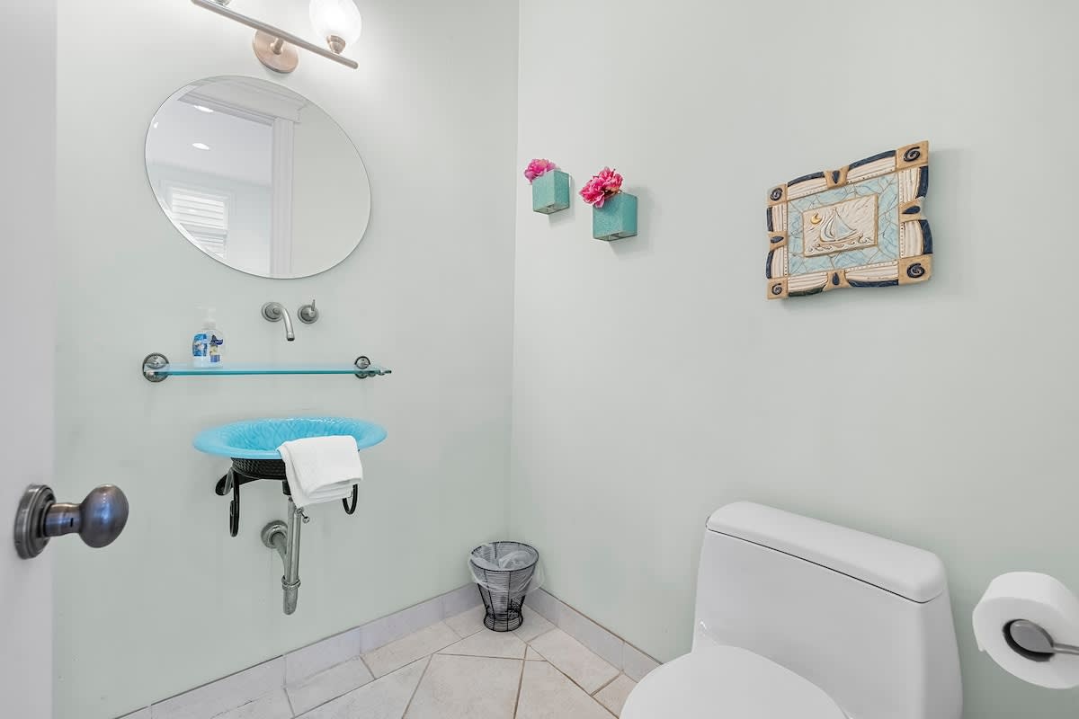 A compact powder room with light green walls, featuring a unique blue glass bowl sink with wall-mounted fixtures, a round mirror, and a white toilet. The floor is tiled, and decorative elements add a touch of color.