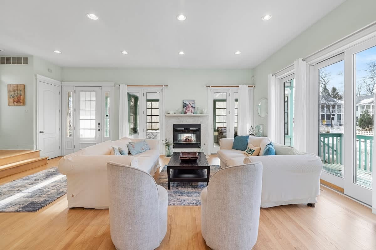 A bright and spacious living room with light green walls, light hardwood floors, two large white slipcovered sofas facing each other, two light gray armchairs, and a dark wood coffee table in front of a white marble fireplace with a gas insert.