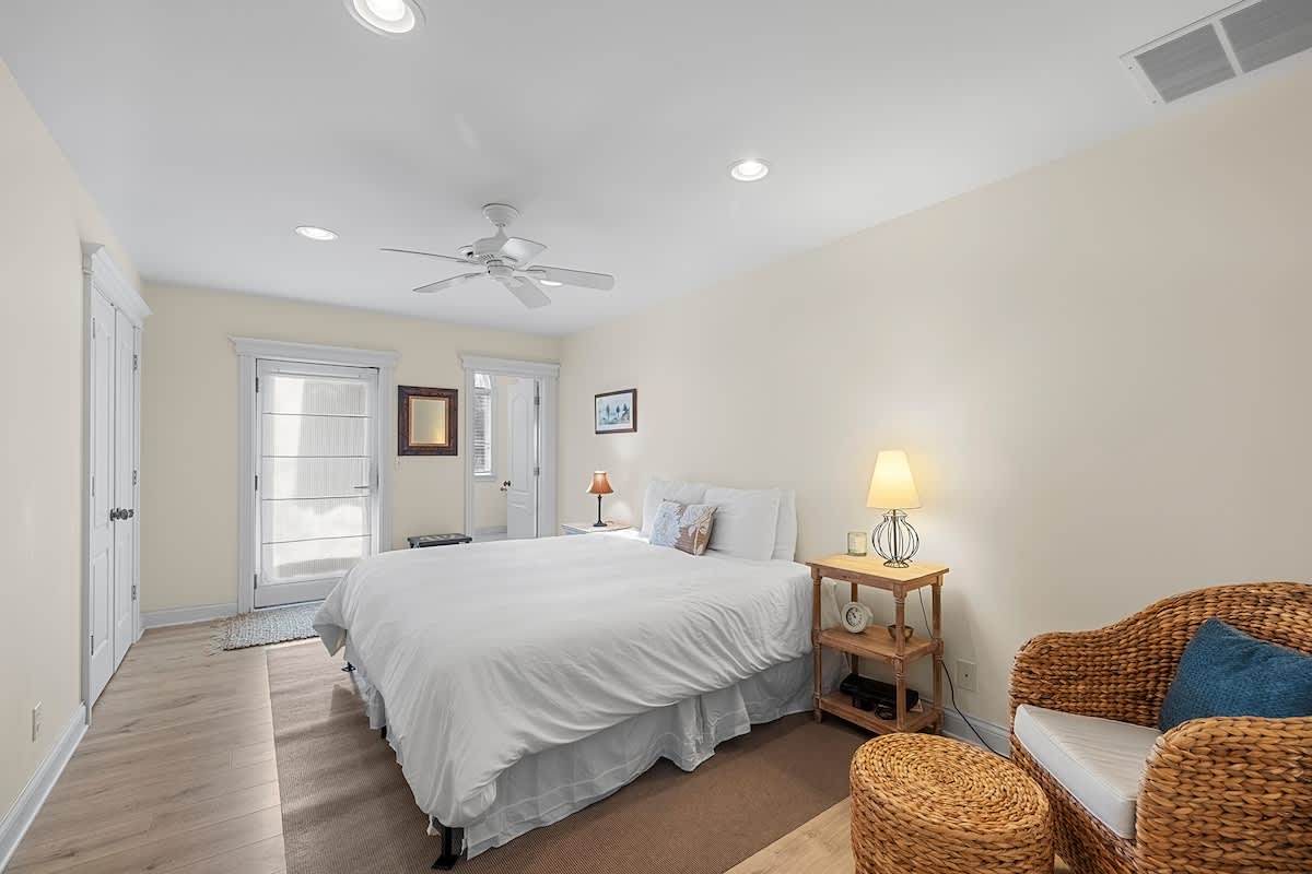 Inviting bedroom featuring a queen bed with white linens, light wood flooring, and a ceiling fan. A wicker chair and side table add a relaxed feel, with a door leading to a balcony.