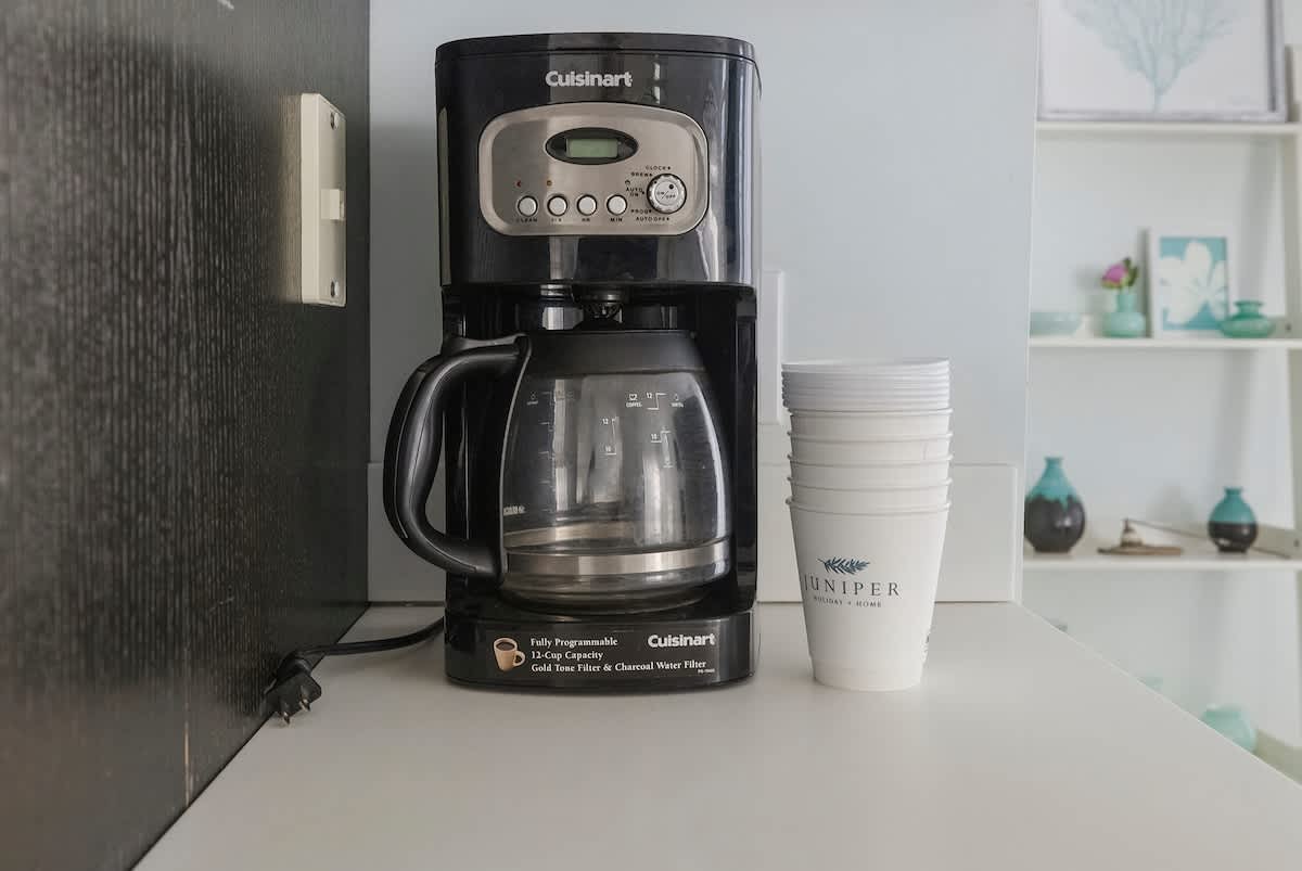 A Cuisinart coffee maker sits on a white countertop next to a stack of disposable cups, with a dark wood panel and white shelving visible in the background.