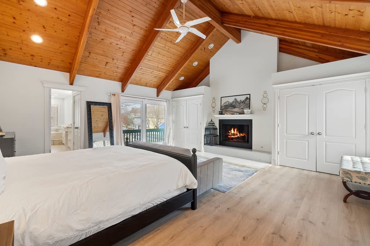 Spacious master bedroom featuring a vaulted wood-paneled ceiling with exposed beams, a cozy fireplace, light wood flooring, and access to a balcony. A large bed with white bedding is centered in the room.
