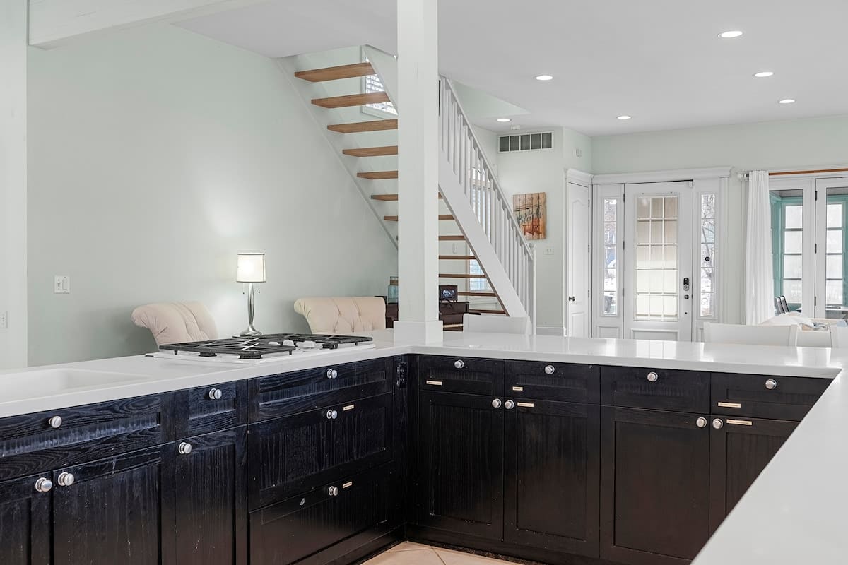 Close-up view of the kitchen island with a gas cooktop and white countertops, showcasing dark wood cabinetry and a tiled floor, with a modern open-tread staircase visible in the background.