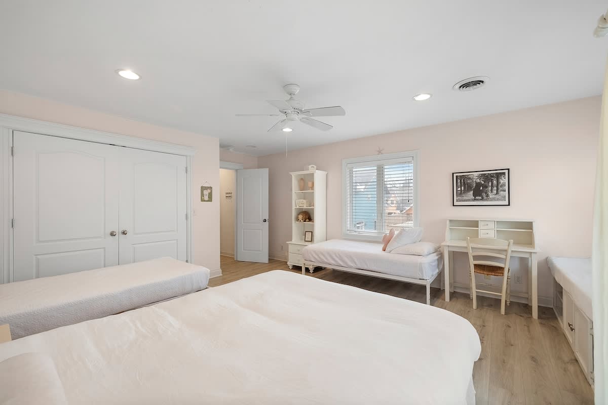 A bright bedroom with light pink walls, featuring two twin beds with white bedding, a third twin bed with a white metal frame, and a white desk with a chair. A ceiling fan and recessed lighting illuminate the space, which has light wood-look flooring.