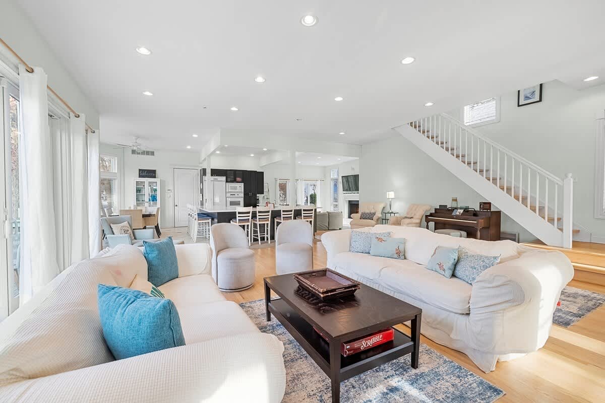 A bright and expansive open-concept living area with light hardwood floors, two large white slipcovered sofas, two light gray armchairs, a dark wood coffee table, and a white staircase leading to the upper floor.