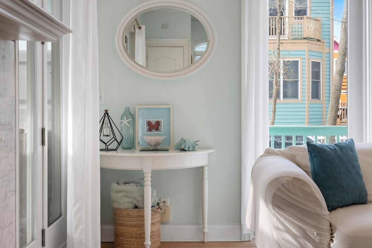 A white oval mirror hangs above a white half-moon console table adorned with coastal-themed decor, including a glass lantern, framed art, and seashells, beside a white curtain and window.