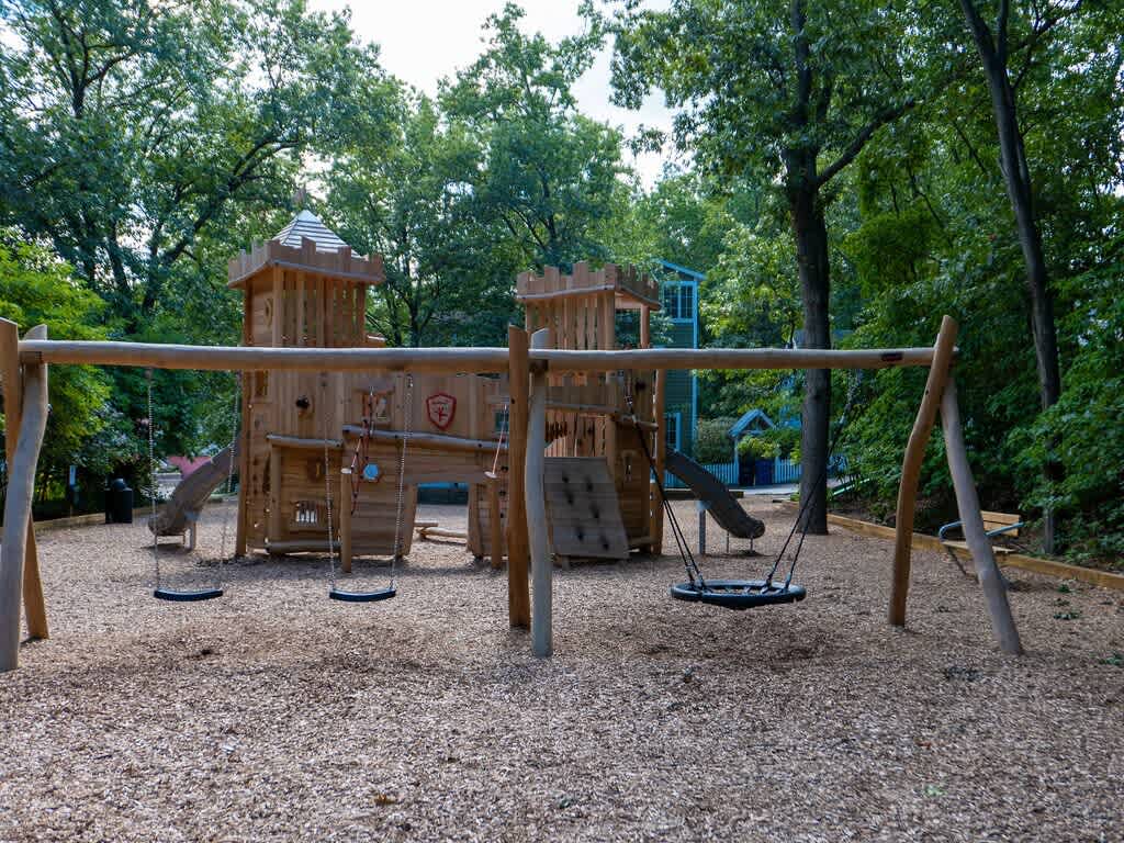 Large wooden playground structure resembling a castle, featuring multiple slides and swings, set on a wood chip surface amidst a dense green forest.
