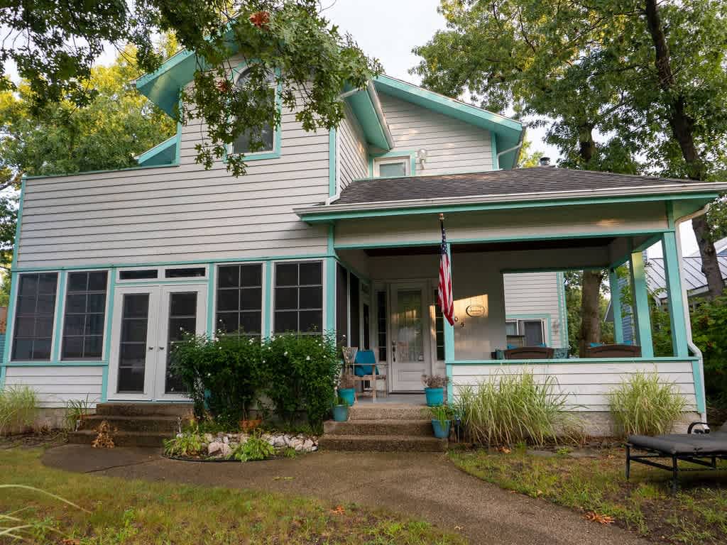 Front exterior of a white house with light blue trim, featuring a screened-in porch with an American flag, a covered front porch, and a stone pathway leading to the entrance.