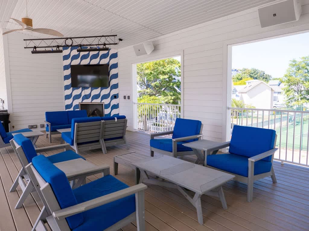 A stylish covered outdoor lounge area features comfortable blue cushioned chairs and sofas arranged around coffee tables, with a large wall-mounted TV above a decorative blue and white patterned fireplace, overlooking a tennis court.