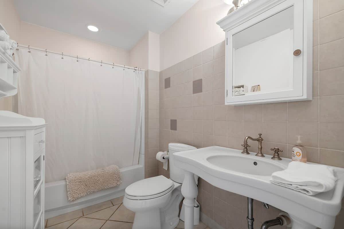 A functional bathroom with light beige tiled walls, a white bathtub with a shower curtain, a white toilet, and a white pedestal sink with brass-toned fixtures. A mirrored medicine cabinet hangs above the sink, and a small rug is on the tiled floor.