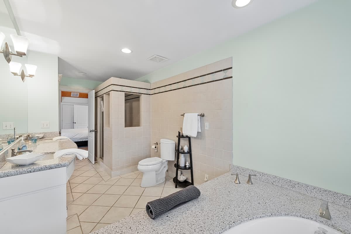 Expansive bathroom with a walk-in shower, toilet, and a long granite double vanity. The room features light green walls and tiled floors and shower walls.