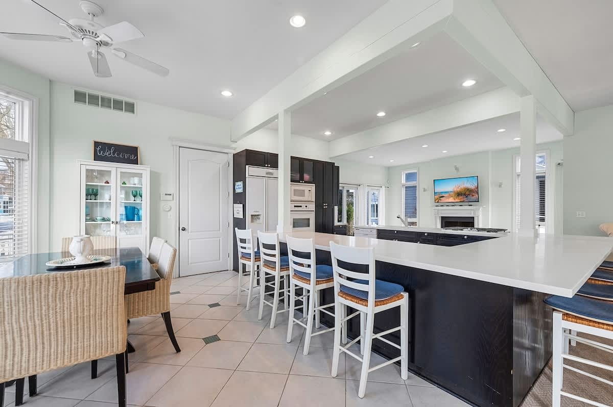 Open-concept kitchen and dining area featuring dark wood cabinetry, white countertops, and a large island with bar seating, adjacent to a dining table with wicker chairs, all under a light green ceiling with a fan.